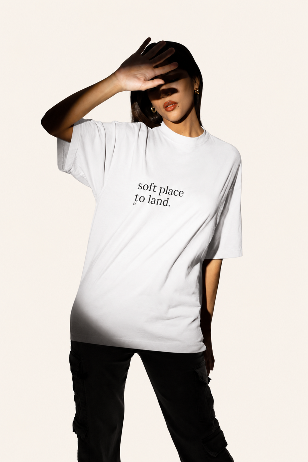 A model wearing an oversized white t-shirt with the black printed text 'soft place to land.' on the chest, paired with black pants, set against a solid off-white background.