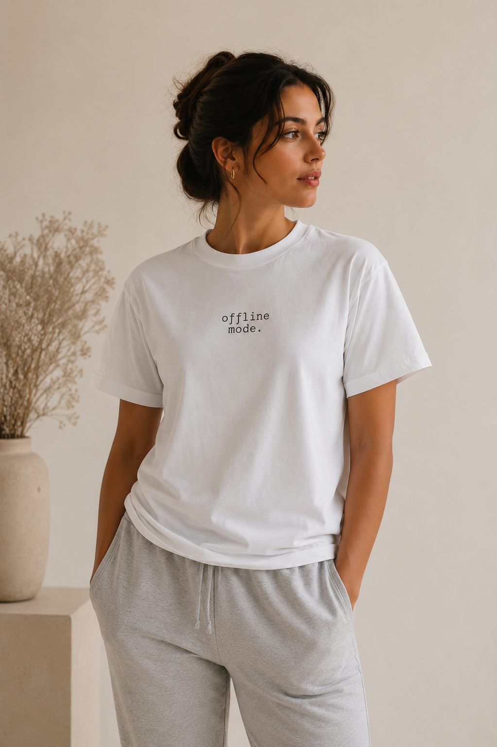 A woman wearing a white crewneck t-shirt with the text 'offline mode.' printed in black on the chest, paired with light gray sweatpants, standing in a minimalist room with a beige wall and a vase of dried flowers.