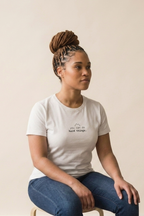 A woman with braided hair in a bun sits wearing a white crewneck t-shirt with a small black graphic on the chest that reads 'the hard things'. She is wearing blue jeans and sitting on a stool against a solid beige background.