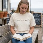 Blonde woman sitting reading wearing Stick to the plan T-shirt in Ivory