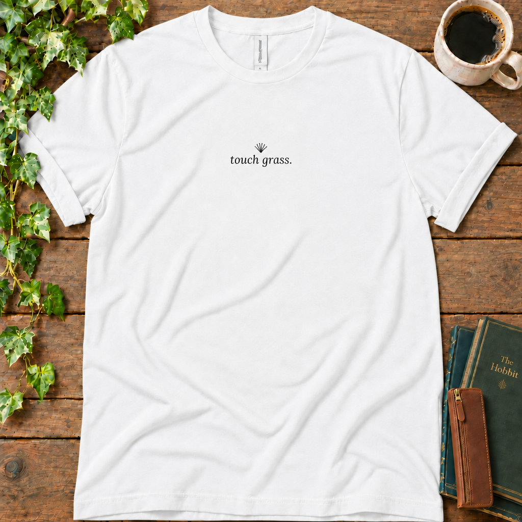 White regular-fit tee printed with touch grass and a small grass sprig, flat lay on rustic wood with ivy, coffee and vintage books