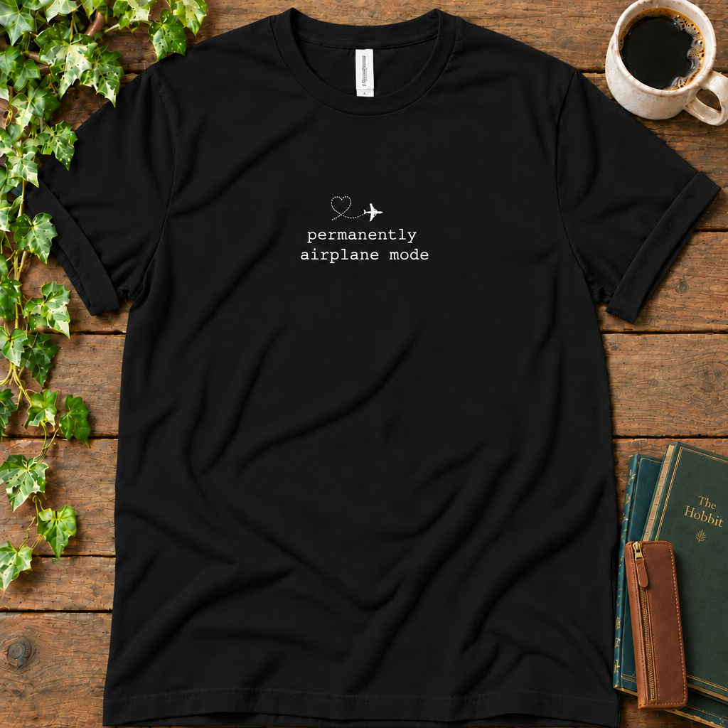 Black regular-fit tee printed with permanently airplane mode and a dotted contrail heart, flat lay on rustic wood with ivy, coffee and vintage books