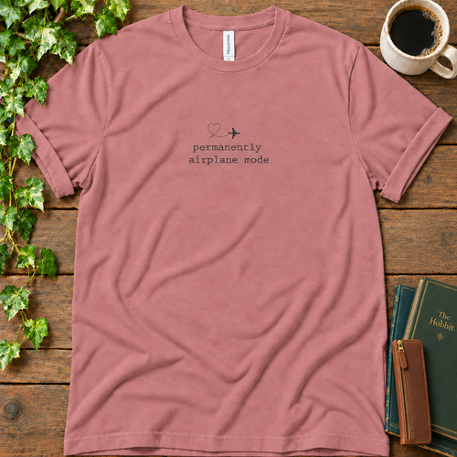 Mauve dusty rose regular-fit tee printed with permanently airplane mode and a dotted contrail heart, flat lay on rustic wood with ivy, coffee and vintage books