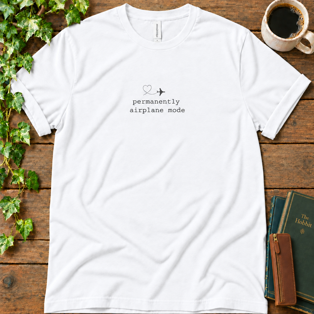 White regular-fit tee printed with permanently airplane mode and a dotted contrail heart, flat lay on rustic wood with ivy, coffee and vintage books