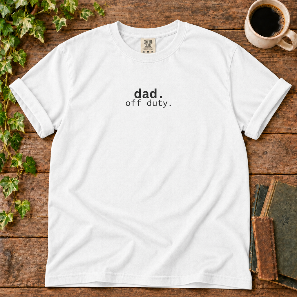 White relaxed-fit tee printed with dad off duty, flat lay on rustic wood with ivy, coffee and vintage books