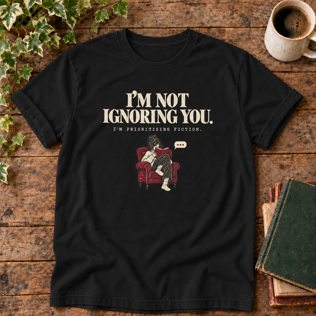 I'm not ignoring you t-shirt in Black, flat lay on dark rustic wood with ivy, coffee mug and green-over-red-and-tan book pile, cream ink