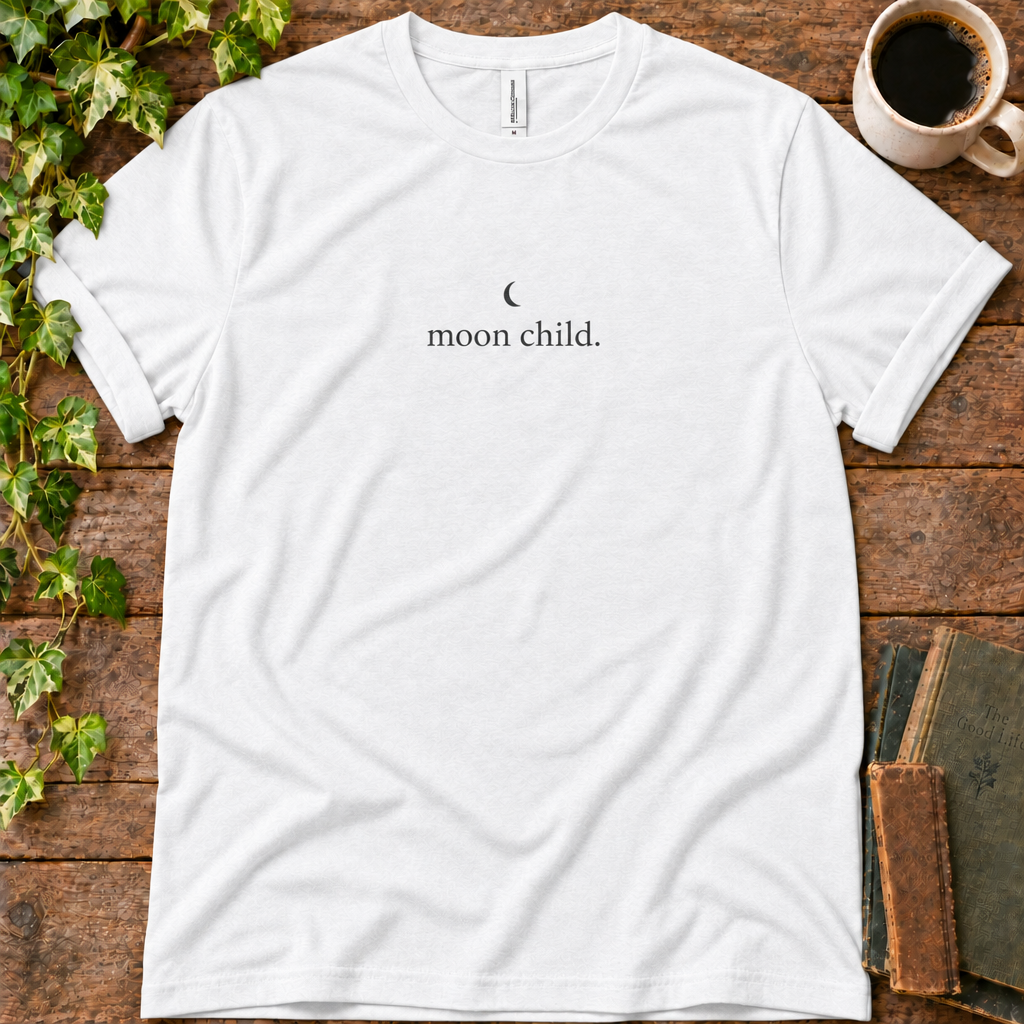 White regular-fit tee printed with moon child, flat lay on rustic wood with ivy, coffee and vintage books