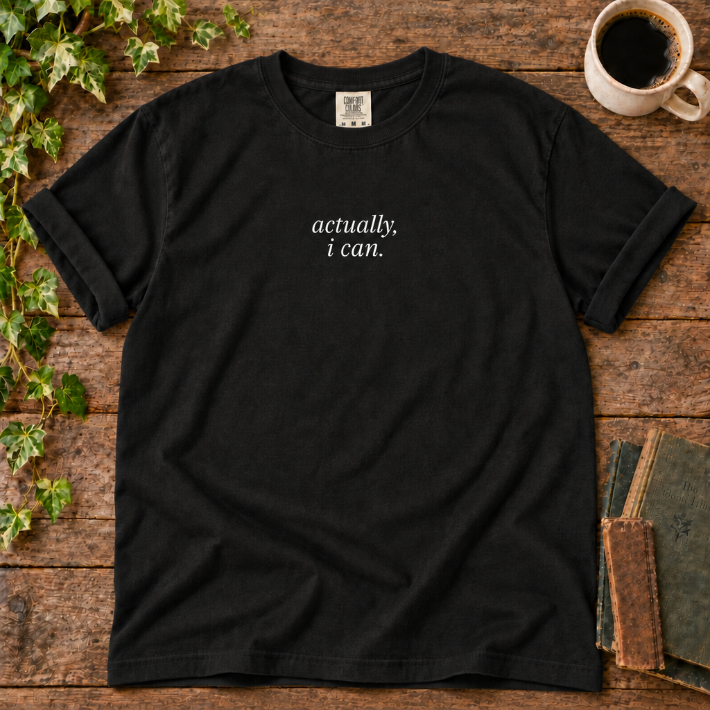Black relaxed-fit tee printed with actually, i can., flat lay on rustic wood with ivy, coffee and vintage books