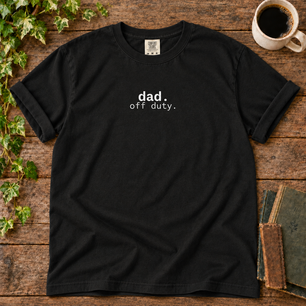Black relaxed-fit tee printed with dad off duty, flat lay on rustic wood with ivy, coffee and vintage books