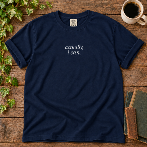 True navy relaxed-fit tee printed with actually, i can., flat lay on rustic wood with ivy, coffee and vintage books