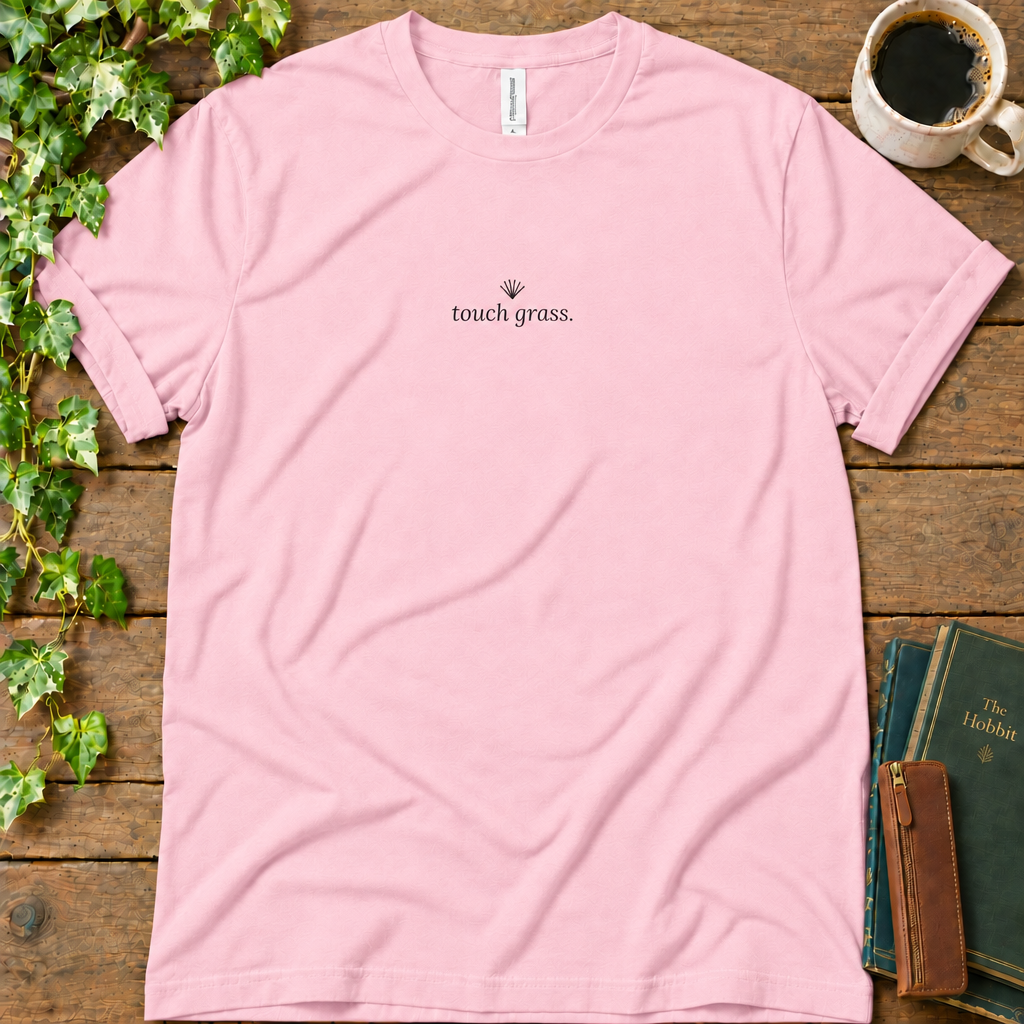 Pink regular-fit tee printed with touch grass and a small grass sprig, flat lay on rustic wood with ivy, coffee and vintage books