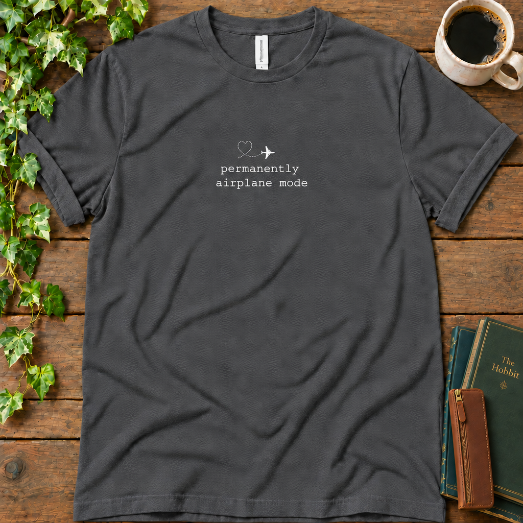 Dark grey heather regular-fit tee printed with permanently airplane mode and a dotted contrail heart, flat lay on rustic wood with ivy, coffee and vintage books