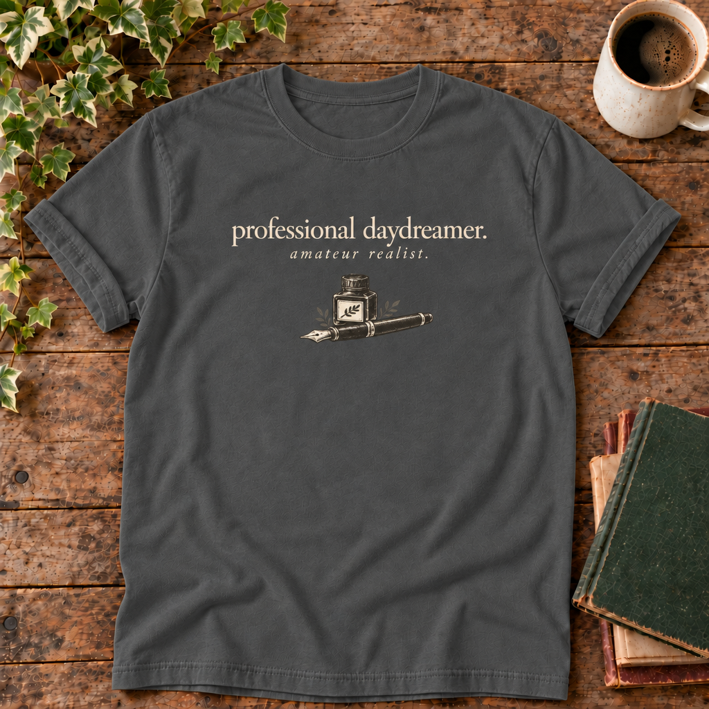 professional daydreamer t-shirt in Pepper, flat lay on dark rustic wood with ivy, coffee mug and green-over-red-and-tan book pile, cream ink