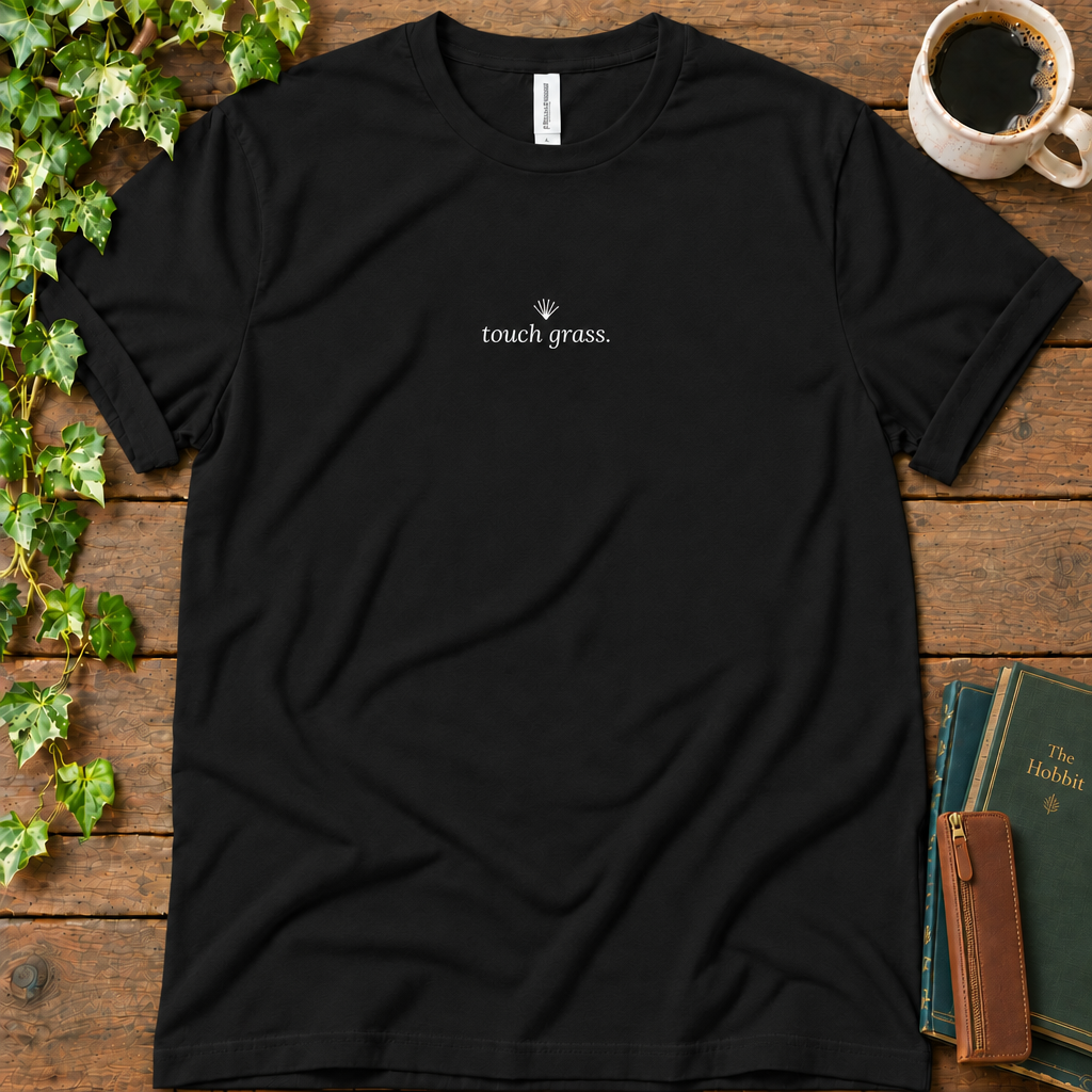 Black regular-fit tee printed with touch grass and a small grass sprig, flat lay on rustic wood with ivy, coffee and vintage books