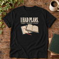 I had plans t-shirt in Black, flat lay on dark rustic wood with ivy, coffee mug and green-over-red-and-tan book pile, cream ink