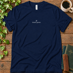 Navy regular-fit tee printed with touch grass and a small grass sprig, flat lay on rustic wood with ivy, coffee and vintage books