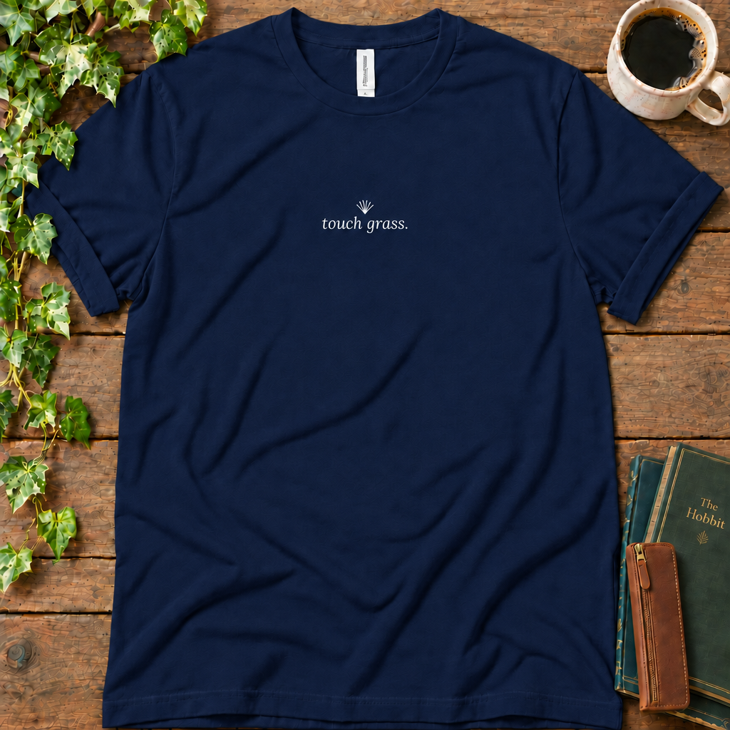 Navy regular-fit tee printed with touch grass and a small grass sprig, flat lay on rustic wood with ivy, coffee and vintage books