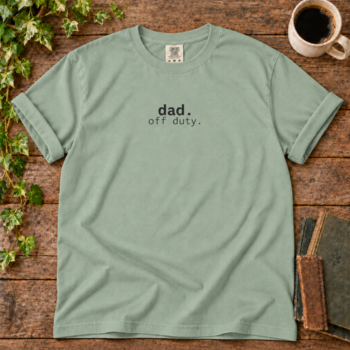 Bay sage green relaxed-fit tee printed with dad off duty, flat lay on rustic wood with ivy, coffee and vintage books