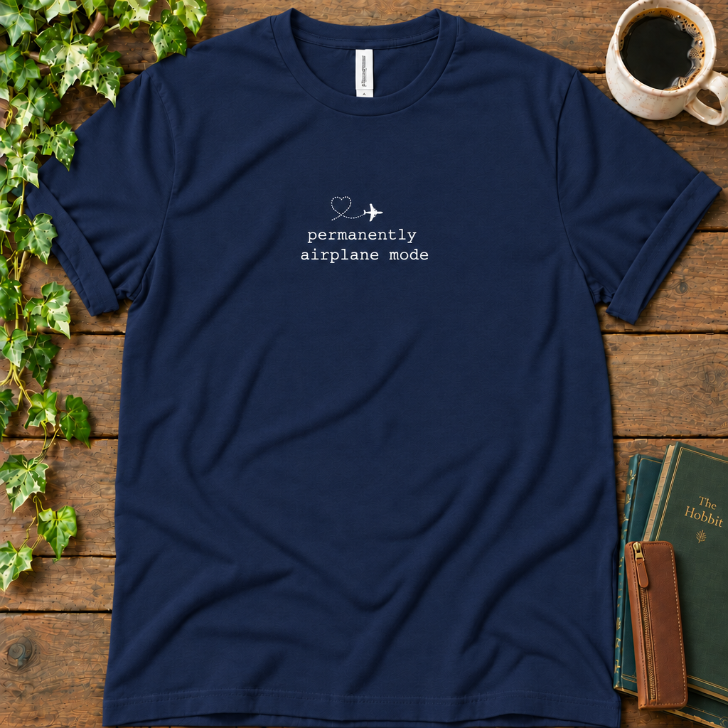 Navy regular-fit tee printed with permanently airplane mode and a dotted contrail heart, flat lay on rustic wood with ivy, coffee and vintage books
