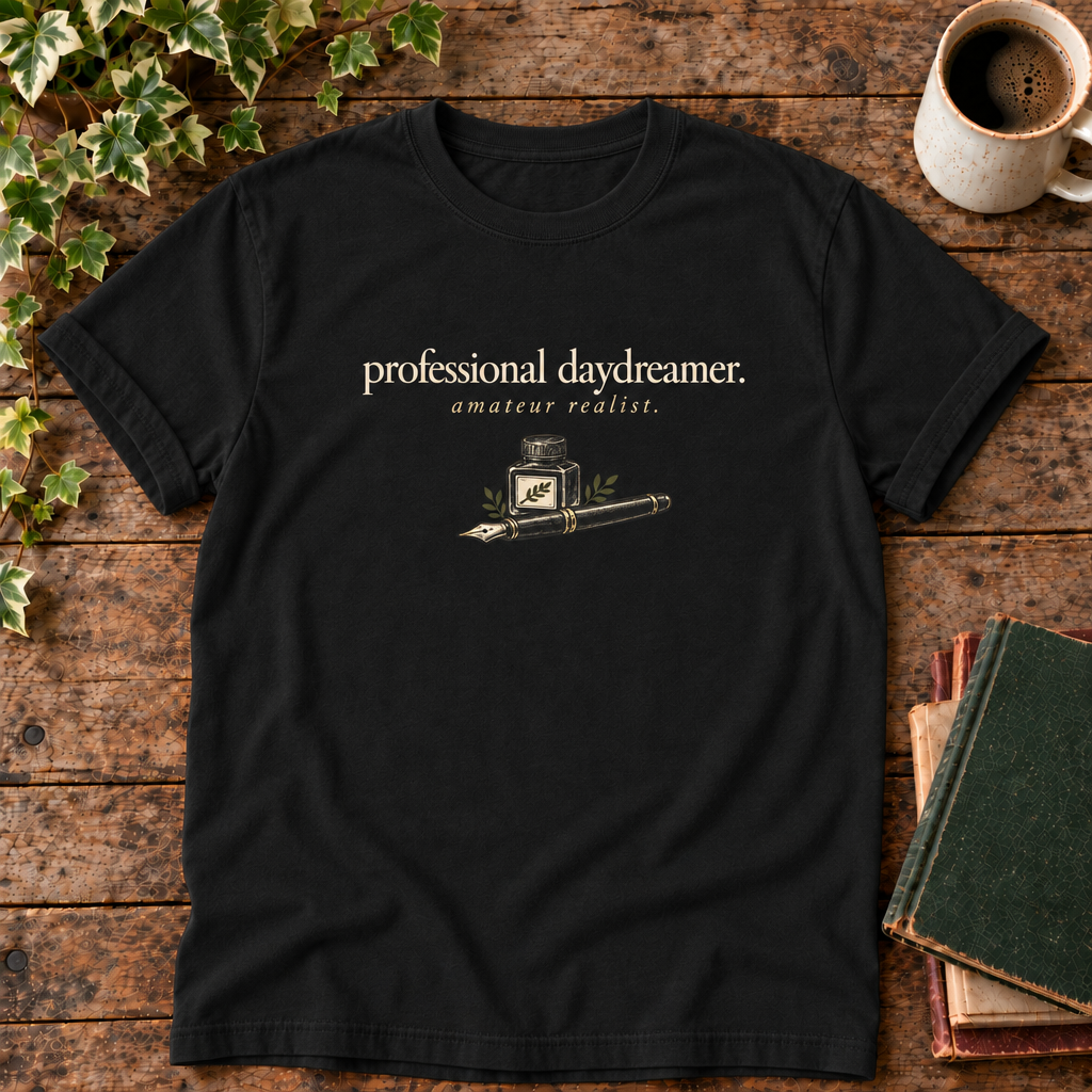 professional daydreamer t-shirt in Black, flat lay on dark rustic wood with ivy, coffee mug and green-over-red-and-tan book pile, cream ink