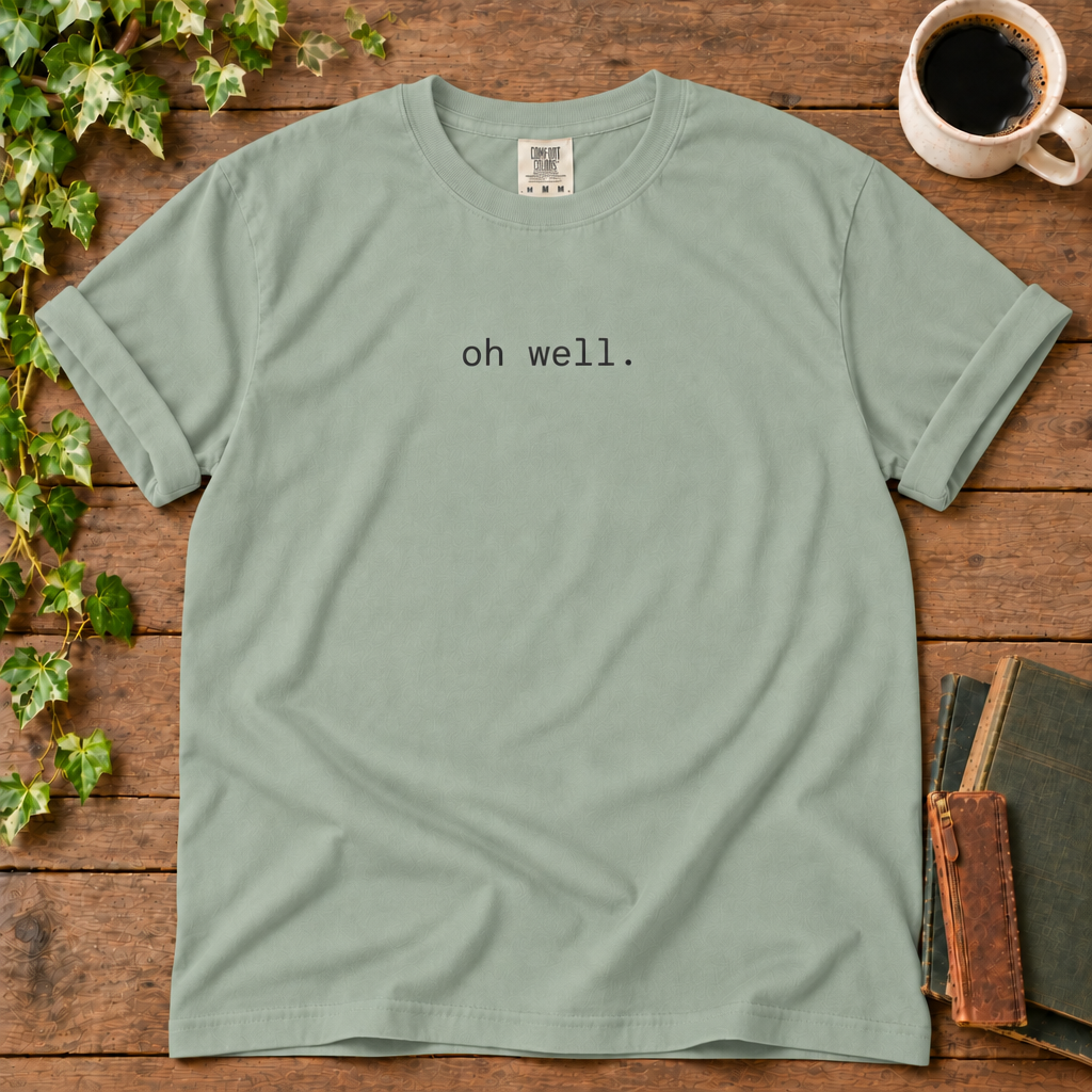 Bay sage green relaxed-fit tee printed with oh well., flat lay on rustic wood with ivy, coffee and vintage books