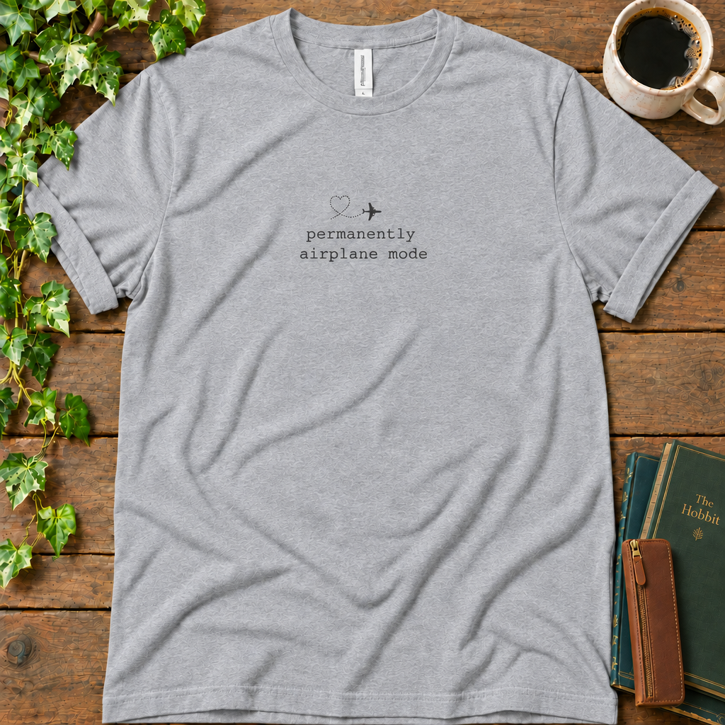 Athletic heather regular-fit tee printed with permanently airplane mode and a dotted contrail heart, flat lay on rustic wood with ivy, coffee and vintage books