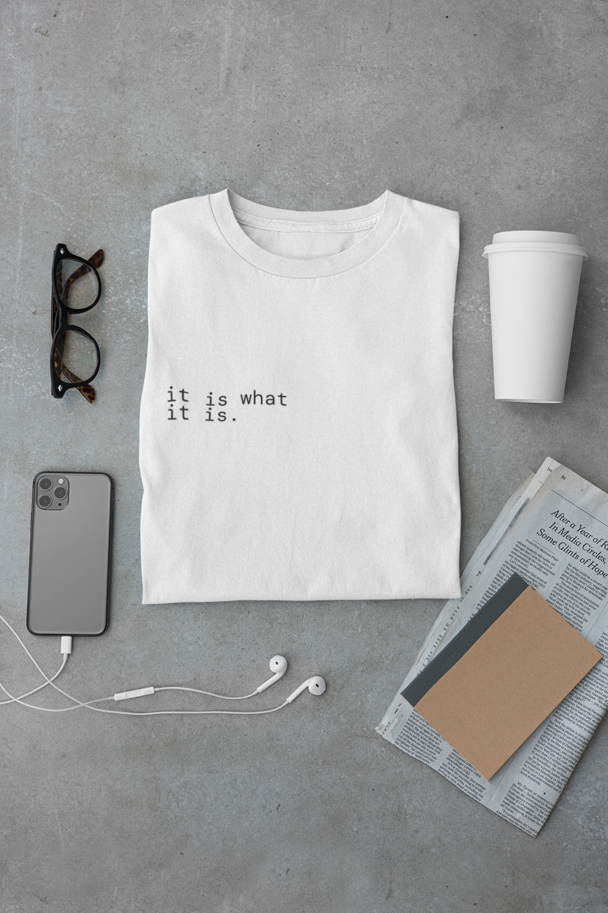 A folded white t-shirt with black typewriter-style text reading 'it is what it is.' is arranged in a flat lay on a gray concrete surface, surrounded by glasses, a smartphone, earbuds, a takeaway coffee cup, and a newspaper with a notebook.