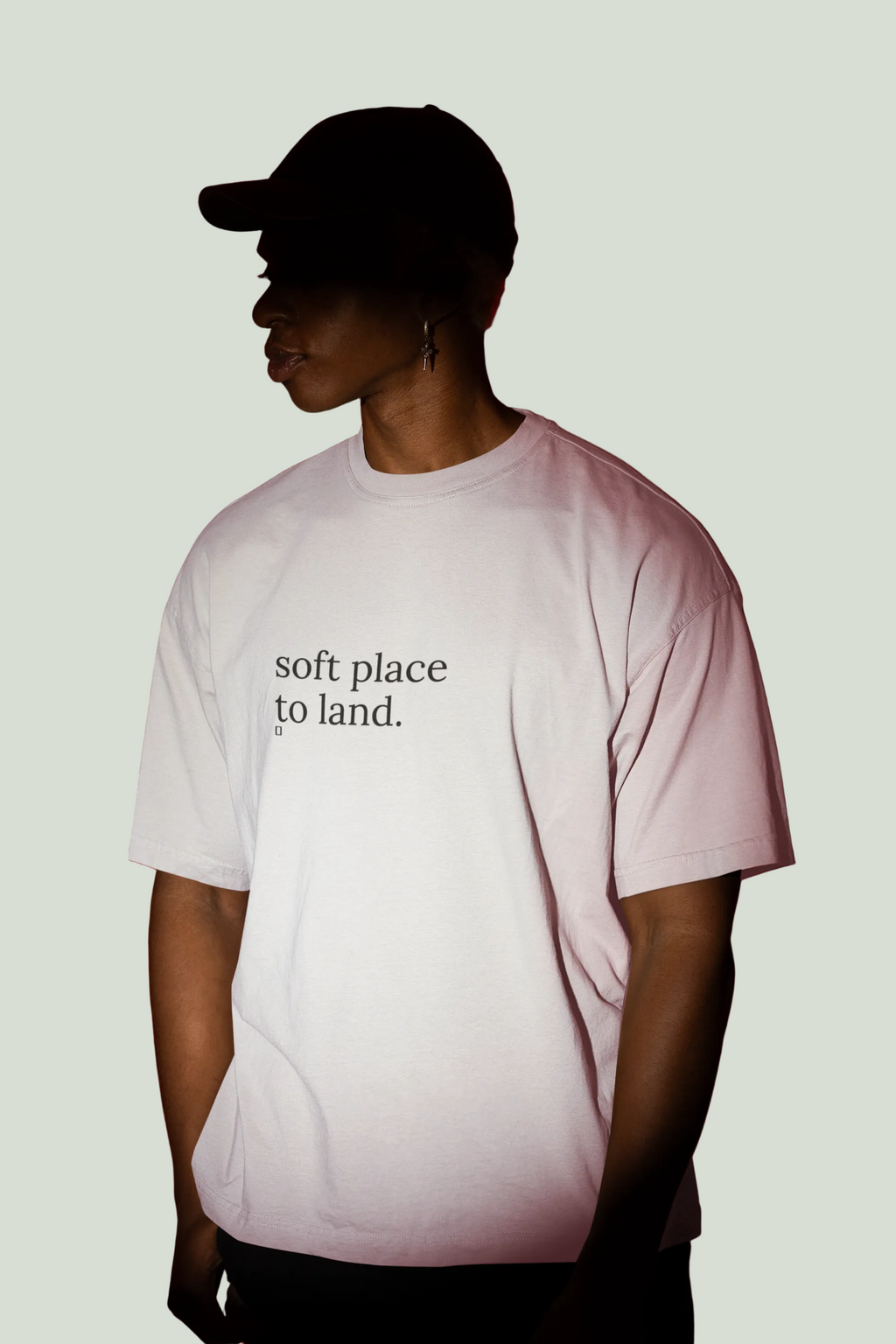 A person in profile wearing a light pink T-shirt with the black text 'soft place to land.' on the chest, paired with a black cap, set against a solid dark red background.
